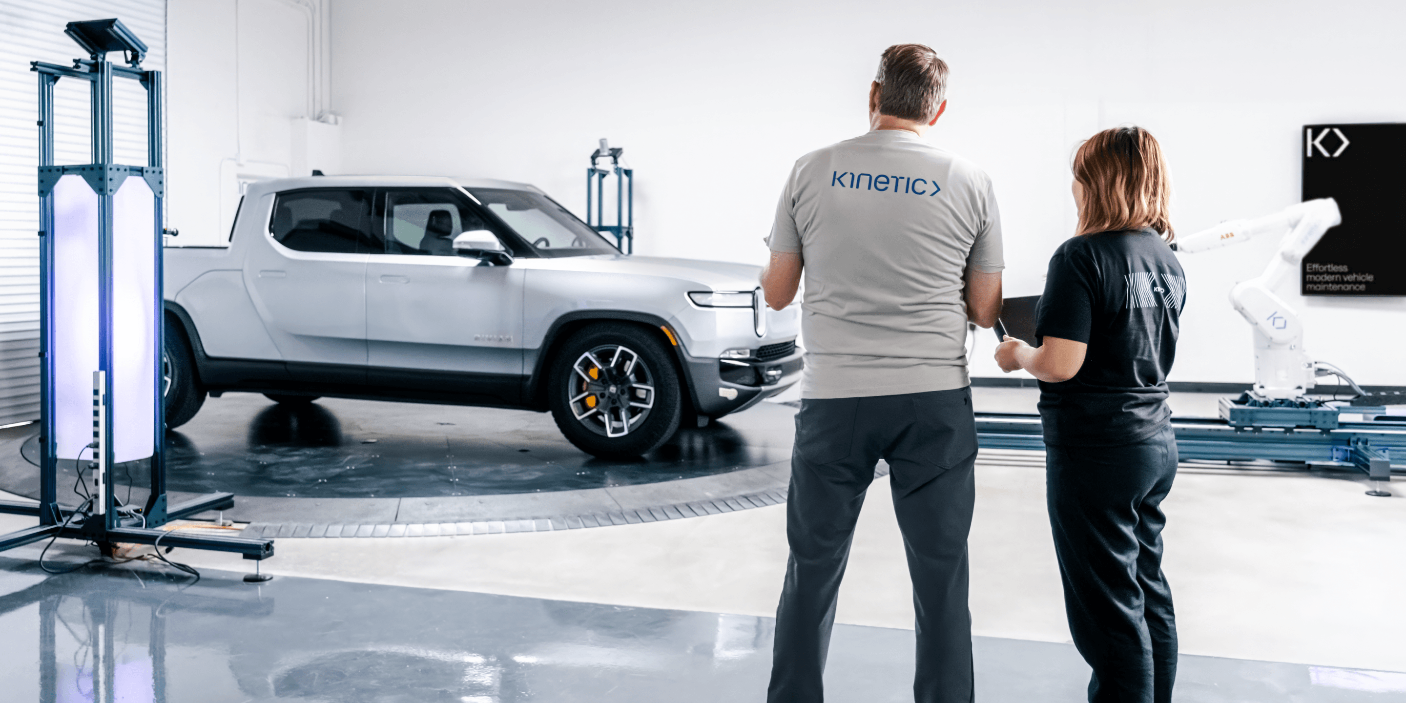 Two Kinetic technicians stand side by side in a Kinetic Hub, assessing an electric silver Rivian pickup truck that sits on a turntable, waiting to be calibrated.