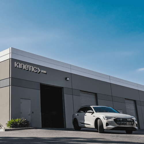 An image showing the exterior of Kinetic’s flagship hub in Orange County, California. A white Audi E-Tron drives out of the garage bay. The Kinetic Hub logo is affixed to the building’s exterior wall.