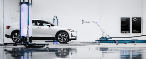 An image showing a Kinetic vehicle maintenance bay. A white Polestar car sits on a turntable, ready to be calibrated by a robotic arm. Two vertical digital screens display a list of cars currently in the service queue, along with the copy line “effortless modern vehicle maintenance.”
